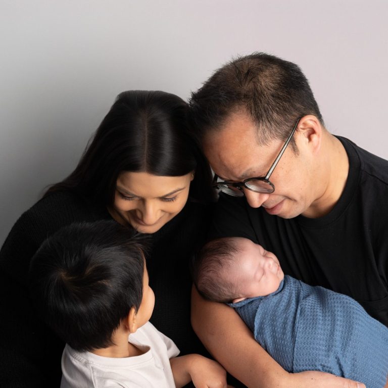 A family of four, smiling and sharing a moment with a newborn and a toddler.