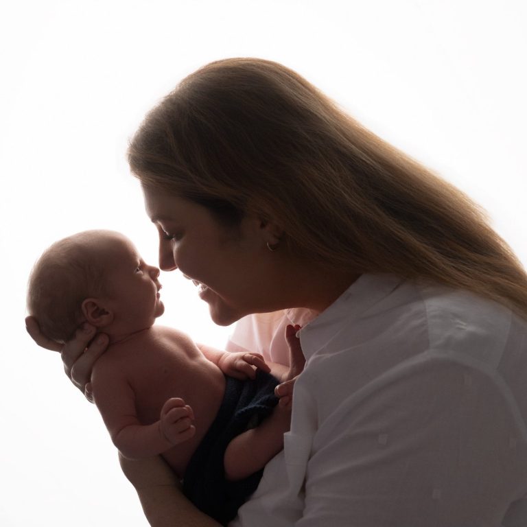 A mother gently holds her newborn, both smiling at each other in soft light.